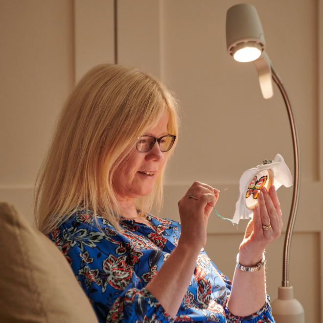 A stitcher threading a needle at her needlework, the work lit by her Serious Readers light
